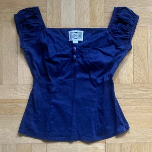 Mörkblå topp från Collectif - Mörkblå 50-tals-inspirerad topp från Collectif. Korta ärmar med rynkade detaljer och två dekorativa knappar framtill! Dragkedja längst ena sidan.