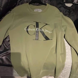 Grön tröja från Calvin Klein - Säljer en snygg grön tröja från Calvin Klein Jeans. Den har en stor CK-logga på framsidan i svart. Tröjan är långärmad och perfekt för en casual look. Passar både till vardags och lite finare tillfällen.