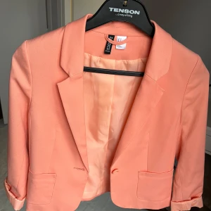 Persikofärgad kavaj från H&M - Säljer en snygg persikofärgad kavaj från H&M i storlek 36. Den har en klassisk krage och är perfekt för både vardag och fest. Kavajen är i mycket bra skick och har en stilren design med långa ärmar och knappar framtill. Passar perfekt till våren och sommaren! 🌸