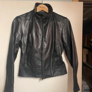 Svart skinnjacka från att. - Säljer en snygg svart skinnjacka från att. i storlek 40. Jackan har en stilren design med dragkedja framtill och långa ärmar. Perfekt för höst och vår, och funkar lika bra till vardags som till fest. 