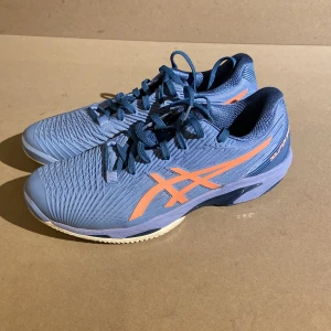Blå och orange Asics Solution Speed FF - Säljer ett par Asics Solution Speed FF i blått med orange detaljer. Skorna har en snygg och sportig design med snörning och en slitstark sula perfekt för tennis. De är i mycket bra skick och redo för nya äventyr på banan!