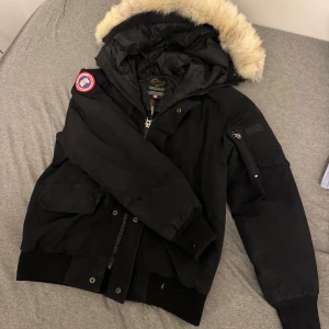 Svart dunjacka från Canada Goose - Säljer en svart dunjacka från Canada Goose med en luva med päls på. Jackan har en dragkedja och knappar framtill, samt flera praktiska fickor. Perfekt för kalla vinterdagar. 