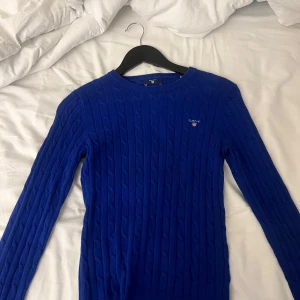 Blå stickad tröja från Gant - Säljer en snygg blå stickad tröja från Gant. Den har ett klassiskt flätmönster och är perfekt för både höst och vinter. Tröjan är långärmad och har en normal passform. Perfekt för att hålla sig varm och stilren! 💙