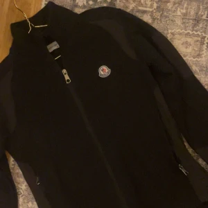 Svart fleecejacka från Moncler - Säljer en svart fleecejacka från Moncler i mycket bra skick. Den har en dragkedja framtill och Monclers logga på bröstet. Perfekt för kyliga höst- och vinterdagar. Jackan är långärmad och har en stilren design.