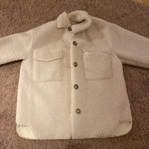 Beige teddyjacka från Zara - Superfin beige teddyjacka från Zara i storlek 11-12 år. Jackan har en mysig och fluffig textur med knappar framtill och två stora fickor. Perfekt för höst och vinter för att hålla sig varm och stilren. 🧥✨