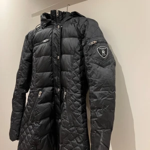 Jacka från Rockandblue - Säljer en snygg svart quiltad jacka från Rockandblue i storlek 36. Jackan har en dragkedja och flera praktiska fickor, inklusive en på ärmen. Perfekt för höst och vinter med sin varma och stiliga design. Huvan ger extra skydd mot kyla. Passar både till vardags och lite finare tillfällen.