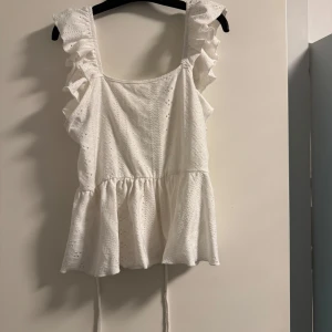 Vit ärmlös topp med volanger - Säljer en superfin vit topp med volanger och öppen rygg. Den är ärmlös och har en söt knytning i ryggen. Perfekt för sommardagar eller en festlig kväll. Toppen har en luftig passform och är i ett lätt material som känns skönt mot huden.
