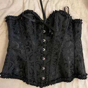 Svart korsett med snörning - Snygg svart korsett med vackert mönster och snörning i ryggen. Den har dekorativa knappar framtill och en söt rosett vid bysten. Perfekt för en festlig kväll eller en speciell tillställning. Passformen är tight och ger en fin siluett. Jag har aldrig använt toppen🥰