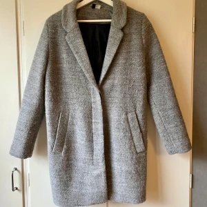 Grå ullkappa - Säljer en stilren grå ullkappa i mycket bra skick. Den har en klassisk design med långa ärmar och två fickor framtill. Perfekt för höst och vinter. Passar både till vardags och lite finare tillfällen. Material: polyester och ull. 