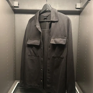 Svart overshirt med knappar - Säljer en stilren svart overshirt i mycket bra skick. Den har långa ärmar och praktiska knappar framtill. Perfekt för höst och vår, och funkar både till vardags och lite mer uppklädda tillfällen. Två fickor på bröstet ger en cool look. Passar perfekt för dig som gillar en enkel men snygg stil.