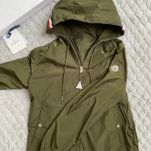Olivgrön vindjacka från Moncler - Säljer en snygg olivgrön vindjacka från Moncler. Jackan har en praktisk huva med röd och vit randdetalj, samt en dragkedja framtill. Perfekt för höst och vår! Den är i mycket bra skick och har Monclers logga på bröstet. Passar perfekt för en stilren look 🍃Priser kan absolut sänkas