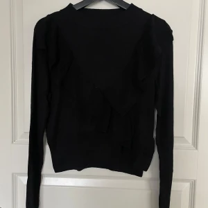 Svart tröja med volangdetaljer - Säljer en stilren svart tröja med volangdetaljer framtill. Tröjan är långärmad och har en normal passform, perfekt för både vardag och fest. Materialet känns mjukt och bekvämt, vilket gör den idealisk för höst och vinter. Ett måste i garderoben för den som älskar klassiska plagg med en twist!