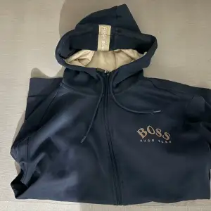 Säljer en mörkblå hoodie från Hugo Boss med dragkedja och broderad logga på bröstet. Den har en skön passform och är perfekt för höst och vår. Tyvärr finns det en liten skada på ena ärmen, men annars i bra skick. Perfekt för en casual look!