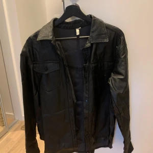 Svart skinnskjorta - Säljer en svart skinnskjorta i bra skick. Den har en klassisk krage och knappar framtill. Perfekt för höst och vår, och ger en cool look till vilken outfit som helst. Skjortan har långa ärmar och två bröstfickor med knappar.