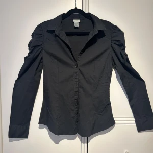 Svart blus med puffärm - Snygg svart blus från H&M i bra skick. Den har långa ärmar med puffiga axlar. Den är v-ringad och har knappar. Ganska figursydd passform. Perfekt för både vardag och fest! 🖤