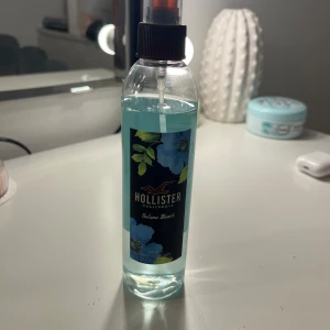 Hollister Solana Beach Body Mist - Fräsch body mist från Hollister med en lätt och uppfriskande doft. Flaskan är genomskinlig med en blå vätska och har en svart etikett med blommönster. Perfekt för en dag på stranden eller när du vill känna dig sommarfräsch.