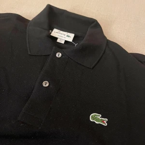 Svart pikétröja från Lacoste - Säljer en klassisk svart pikétröja från Lacoste i mycket bra skick. Tröjan har en broderad krokodil på bröstet och knappar framtill. Perfekt för både vardag och fest!