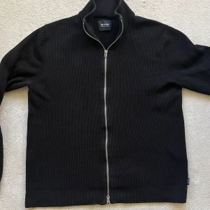 Full Zip från Only & Sons - Säljer en stilren svart ribbad tröja från Only & Sons. Den har en dragkedja framtill och är perfekt för både vardag och fest. Tröjan är långärmad och har en normal passform. Perfekt för höst och vinter!