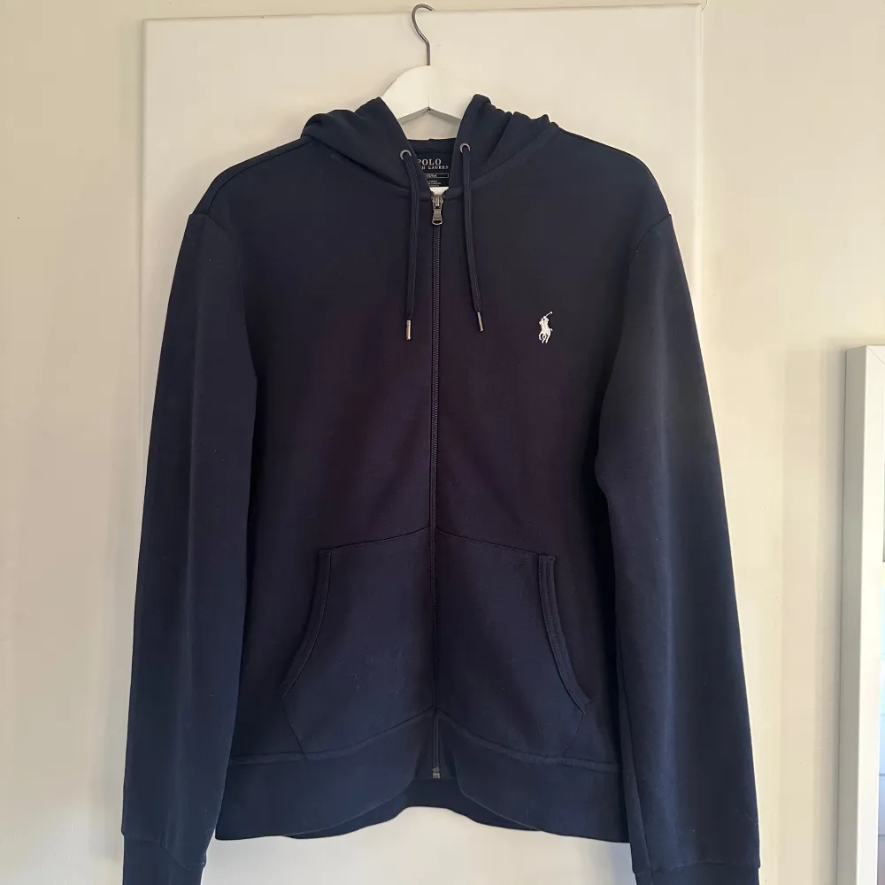 Säljer en snygg mörkblå hoodie från Polo Ralph Lauren i storlek M. Den har en dragkedja framtill och en klassisk logga broderad på bröstet. Perfekt för alla årstider, med en skön passform och praktiska fickor. Finns inga tecken på användning, som nu utan defekter. Ordinarie pris 2295kr (modell: 30/1 DOUBLE KNT CVC-LSL-KNT) Herrmodell men funkar absolut som unisex!. Tröjor & Koftor.