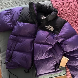 Vinterjacka från The North Face - Säljer en supermysig HELT OANVÄND lila dunjacka från The North Face i modellen 1996 Retro Nuptse. Den är perfekt för kalla dagar med sin pufferstil och har en dragkedja framtill. Jackan är i mycket bra skick och har en snygg svart detalj upptill. Perfekt för vintern! 💜 Orginalpris: 3000kr Skicka bud om ni vill 
