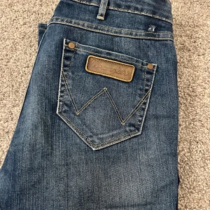 Blå bootcutjeans från Wrangler - Säljer ett par klassiska blå jeans från Wrangler i modellen Betty. Hål upptill. De är i bra skick och har en snygg, enkel design med en bakficka med logotyp. Perfekta för vardagsbruk och passar till det mesta. 