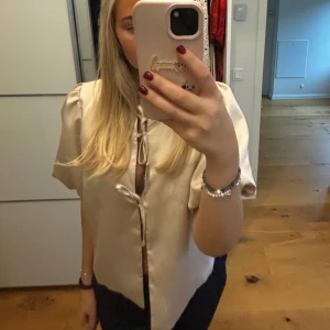 Beige satinblus med knytning - Säljer en elegant beige blus i satin med korta ärmar och knytning framtill. Perfekt för både vardag och festliga tillfällen. Blusen har en lös passform och ger en lyxig känsla. Passar bra till både jeans och kjol. Helt ny med lapp kvar.