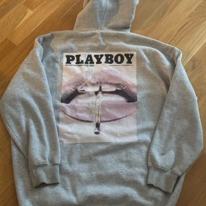 Grå hoodie med Playboy-tryck - Säljer en grå hoodie med stort Playboy-tryck på ryggen. Den har en klassisk känguruficka och en bekväm huva. Perfekt för en avslappnad stil och att hålla sig varm under kyliga dagar. Passar både till vardags och chillkvällar.