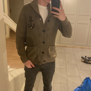 Olivgrön stickad kofta från G-Star RAW - Säljer en fet grön stickad kofta från G-Star RAW i storlek L. Den har en cool ankardetalj på bröstet och praktiska fickor framtill. Perfekt för höst och vinter, med en skön och varm känsla. Passar både till vardags och lite mer avslappnade tillfälle
