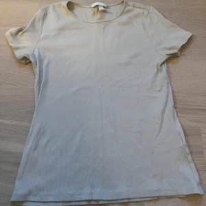 Beige ribbad t-shirt från H&M - Säljer en stilren beige ribbad t-shirt från H&M i storlek M. Perfekt för vardagsbruk med sin enkla design och bekväma passform. Kortärmad och gjord i ett mjukt material som känns skönt mot huden. Passar till alla tillfällen!