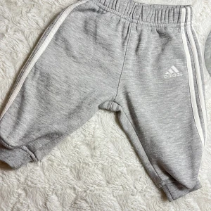 Grå mjukisbyxor från Adidas - Säljer ett par grå mjukisbyxor från Adidas i barnstorlek 68. Byxorna har en elastisk midja och vita ränder längs sidorna.  Adidas-loggan är broderad på framsidan. Superbekväma och i bra skick!