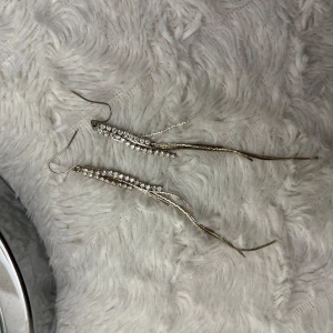 Glittriga silverörhängen med strass - Säljer ett par eleganta och glittriga silverörhängen med strassdetaljer. De har långa, smala hängen som ger en snygg och festlig look. Perfekta för en utekväll eller speciella tillfällen. Örhängena är i mycket bra skick och redo att bäras för att lysa upp din outfit! Dessa är aldrig använda 🤩 