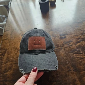 Svart Acne-keps med läderdetalj - Säljer en svart keps med en cool läderdetalj framtill. Kepsen har en sliten look med fransar vid skärmen, vilket ger den en avslappnad stil. Perfekt för vardagsbruk eller som en statement-accessoar.