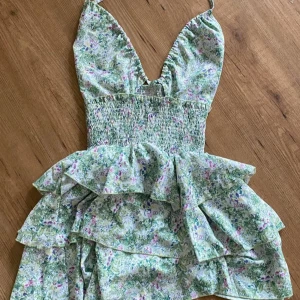 Grön blommig klänning med volanger - Säljer en superfin grön klänning med blommigt mönster. Den har smala axelband och en v-ringad urringning. Klänningen är kort och har flera lager av volanger som ger en härlig sommarkänsla. Perfekt för sommarens alla fester!