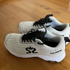 Vita sneakers från Salming - Säljer ett par vita Salming sneakers i nyskick. De har en stilren design med svarta detaljer och snörning. Perfekta för träning eller vardagsbruk. Skorna har en bekväm passform och är tillverkade i syntetmaterial. Köpt för 1885 säljer nu för 999