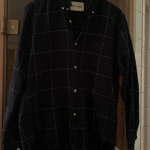 Svart rutig skjorta från Our Legacy - Säljer en stilren svart skjorta från Our Legacy med ett diskret rutmönster i vitt. Skjortan är i gott skick och Storlek 48