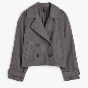 Grå kavaj med knappar - Säljer en snygg grå kavaj med dubbelknäppning och breda slag. Den är croppad och har en loose passform, perfekt för höst och vår. Kavajen har långa ärmar med dekorativa knappar vid ärmsluten. Materialet känns som ull och ger en stilren look.