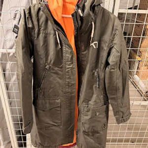 Grön parkajacka från Kari Traa - Säljer en snygg grön parkajacka från Kari Traa. Den har en cool orange insida och är perfekt för höst och vår. Jackan har långa ärmar och praktiska dragkedjor på fickorna. Dessutom finns det en justerbar bälte i midjan för en bättre passform. Perfek