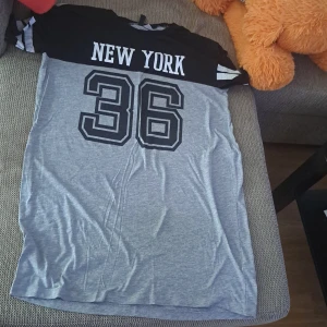 Grå och svart t-shirt med New York-tryck - Säljer en cool t-shirt i grått och svart med texten 'New York' och siffran '36' tryckt på framsidan. T-shirten har korta ärmar med vita ränder och är perfekt för en avslappnad stil. Passar bra till jeans eller shorts för en sportig look.