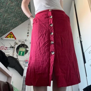 Röd kjol med knappar - Säljer en superfin röd kjol med knappar hela vägen ner. Den har en härlig färg och är perfekt för både vardag och fest. Kjolen är i ett skönt material och har en liten slits framtill för extra rörelsefrihet. Passar perfekt till sommardagar eller en mysig höstkväll.