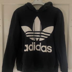 Svart hoodie från Adidas - Säljer en svart hoodie från Adidas med den klassiska loggan i vitt på framsidan. Den är i bra skick och perfekt för både vardag och träning. Mjuk och bekväm med långa ärmar och en praktisk huva. Passar perfekt till höst och vinter!