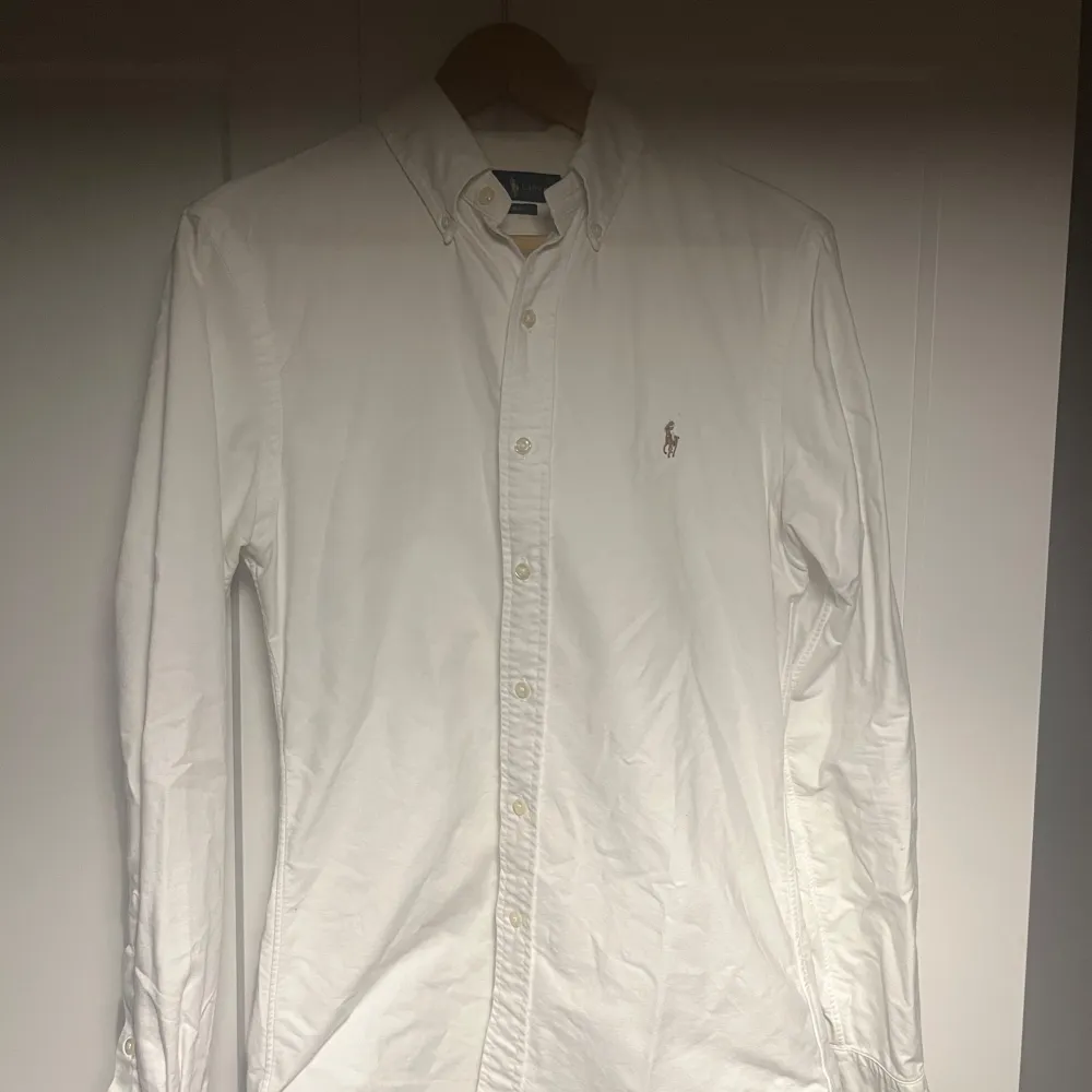 Säljer en stilren vit skjorta från Ralph Lauren i slim fit. Skjortan har långa ärmar och klassiska knappar framtill. Perfekt för både vardag och festliga tillfällen. Den är i bra skick och redo för en ny ägare!. Skjortor.