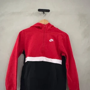 Röd och svart hoodie från Nike - Säljer en snygg röd och svart hoodie från Nike. Den har en halv dragkedja framtill och en vit rand som delar färgerna. Perfekt för både träning och vardag. Den är i bra skick och superbekväm att ha på sig. Passar perfekt till höst och vår!