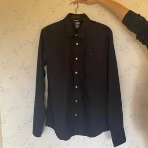 Svart skjorta från Hilfiger Denim - Snygg svart skjorta från Hilfiger Denim i storlek XL. Den har långa ärmar och klassiska knappar framtill. Perfekt för både vardag och fest! Skjortan är i bra skick och har en liten broderad logga på bröstet. Passar perfekt till jeans eller chinos för en stilren look.