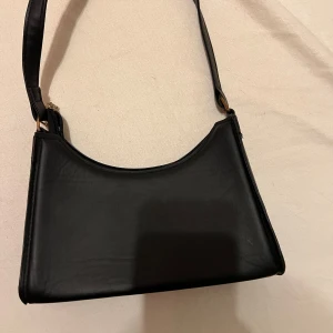 Svart axelväska i skinn - Snygg svart axelväska i skinn med dragkedja. Perfekt för både vardag och fest. Väskan har en stilren design och är i mycket bra skick. En klassisk accessoar som passar till allt! Säljs för 60