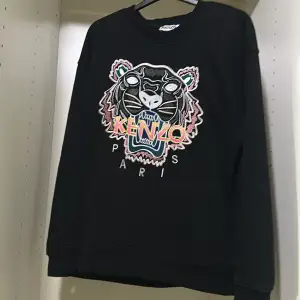 Säljer en svart sweatshirt från Kenzo med ett coolt broderat tigerhuvud och färgglatt Kenzo Paris-tryck på framsidan. Tröjan har långa ärmar och är perfekt för en avslappnad stil. Passar både till vardags och festliga tillfällen.