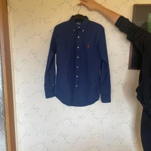 Blå skjorta från Polo Ralph Lauren - Säljer en snygg blå skjorta från Polo Ralph Lauren i storlek S. Skjortan har långa ärmar och klassiska knappar framtill. Perfekt för både vardag och festliga tillfällen. Den är i bra skick och redo för en ny ägare!