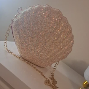 Glittrig snäckformad väska med kedja - Säljer en superfin snäckformad väska med glittrigt material och en elegant kedja. Perfekt för festliga tillfällen! Väskan har en dragkedja och är i en skimrande beige färg med en härlig röd insida. Den är både söt och praktisk för att bära med sig det viktigaste.