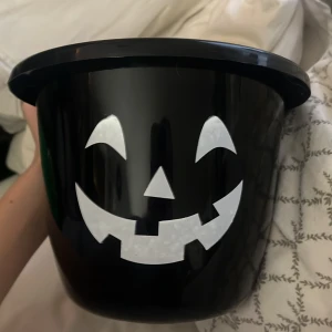 Svart Halloween-hink med ansikte - En svart hink med ett vitt, glatt ansikte som påminner om en pumpa. Perfekt för Halloween-firande och trick-or-treat! Hinken har ett handtag för enkel bärning.