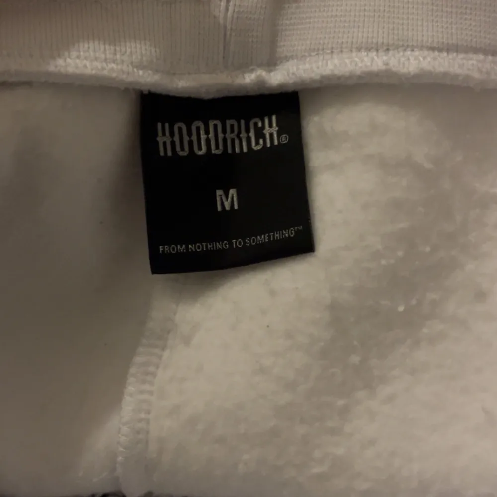 Tjena, jag säljer ett par vita mjukisbyxor från Hoodrich i storlek M. Byxorna har en cool svart och röd logga på framsidan och en ficka på sidan. Perfekta för en avslappnad stil eller chill dagar hemma. De är i bra skick och är skit bekväma!. Farkut & Housut.