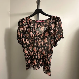 Blommig blus - Blommig blus i svart och rosa. Den har korta ärmar med volangdetaljer. Perfekt för sommardagar eller en festlig kväll. Från Indiska i storlek L!